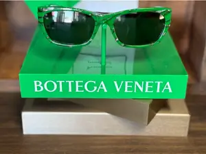 A pair of Bottega Veneta sunglasses in green frames displayed on a green box with the brand name on it.