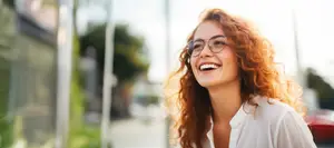 A woman with curly red hair and glasses smiles on a street with blurred background