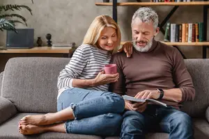 An older man and woman are sitting on a couch, smiling and holding a cup and a book, respectively