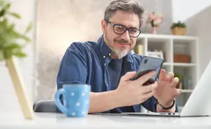 A man in glasses sits at a desk using a mobile phone and a laptop with a coffee mug nearby.