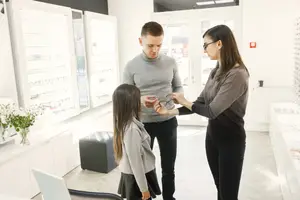 Two adults and a young girl are standing in front of a table, one of the adults is holding a pair of glasses in their hands, while the other adult is looking at the glasses, and the young girl is looking at the glasses as well. Behind them, there is a shelf with a white vase and a flower, a window with a view of a building, and a fire extinguisher.