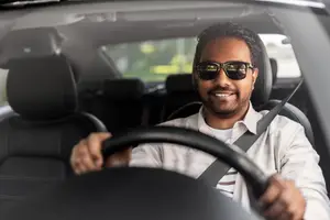 A man wearing sunglasses and a seatbelt smiles while driving a car with his left hand on the steering wheel and his right hand on the seat.