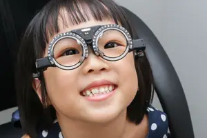 Image of a smiling young girl with black hair wearing eyeglasses with a black frame and silver lenses sitting on a black chair in a room