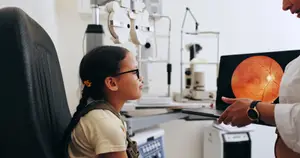 A girl with glasses is looking at an eye exam machine while a woman in a white coat holds a laptop showing an image of an eye.