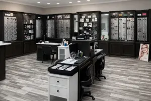 Inside a well-lit office space, there are several desks and chairs arranged with various items on them. On the left side, there are wooden cabinets filled with eyeglass frames displayed on glass shelves. On the right side, there is a picture of a woman with glasses. The floor is covered with gray tiles, and the room has a white wall and ceiling.