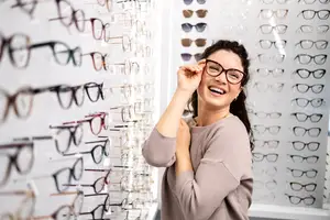 A woman in front of a wall of glasses smiling while holding a pair of glasses to her face