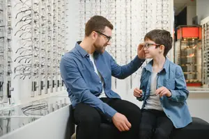 A man and a boy are sitting on a bench and looking at glasses in a store.