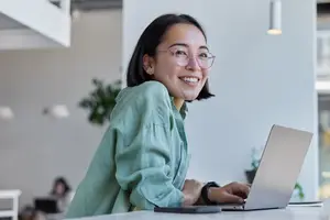 Asian woman wearing glasses sitting at a desk with a laptop and smiling