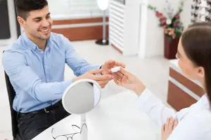 A man and a woman are in a room, the man is holding a contact lens and the woman is looking at him while holding his hand