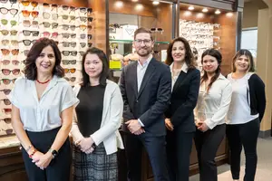 Six individuals dressed in professional attire are standing together in front of a glass display case filled with sunglasses.