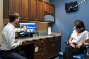 Man pointing to eye scan on computer monitor while woman sits nearby in blue chair
