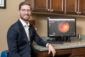 A man is sitting on a chair in front of a desk with a computer monitor, keyboard, and mouse. He is wearing glasses and a watch, smiling, and leaning on the desk.