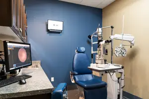 A room with medical equipment including an eye examination chair, an ophthalmoscope, and a computer monitor displaying an eye image.