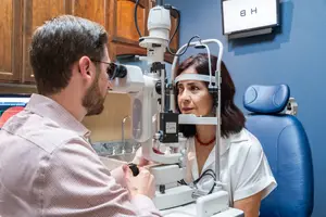 A man in glasses is using an ophthalmoscope to examine a woman's eyes in an office