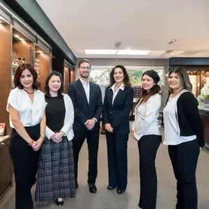 A group of people, possibly employees, standing in a room with cabinets, a flower vase, and windows. They are smiling and posing for a photo. One man is holding a cell phone, and the woman on the left is wearing a necklace and bracelets.
