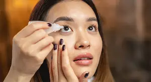 A woman applying eye drops with a serious expression, holding a bottle in one hand and touching her eye with the other