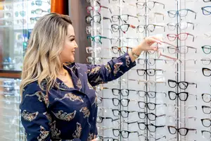 A woman with blonde hair is standing in front of a display of glasses in a shop, pointing to a pair of glasses with her right hand.