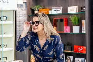 A woman wearing glasses is standing in front of a glass shelf with several items displayed.
