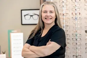 A woman with blonde hair is smiling and posing for a photo in front of a display of glasses.
