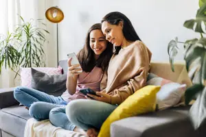 Two women sitting on a couch, one looking at her phone and the other smiling and looking at the phone of the woman beside her. They are surrounded by plants and pillows on the couch.