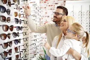 A man, a woman, and a young girl are looking at a wall of sunglasses in a store.