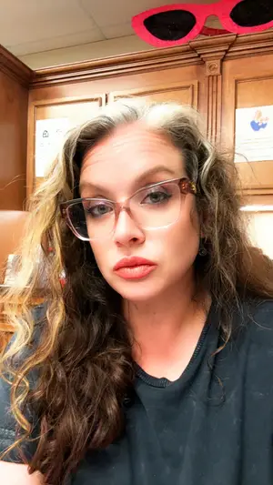 A woman with curly hair and glasses wearing a black t-shirt is sitting in a wooden room.