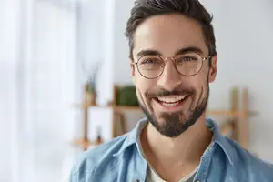 A man with a beard and glasses smiles in a room with wooden shelves and a white curtain.