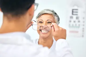 A woman wearing glasses is smiling and looking at a man in front of her, who is also wearing glasses and holding the glasses of the woman.