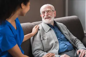 An elderly man sitting on a couch with a caregiver, both smiling at each other.