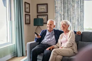 Two elderly people sitting on a couch, smiling while watching TV using a remote control.