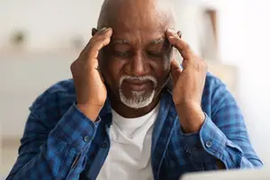 A close-up of a bald older man in a blue shirt sitting with his head in his hands, possibly in a living room.