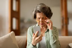 An elderly woman with gray hair wearing glasses sits on a couch holding a phone.