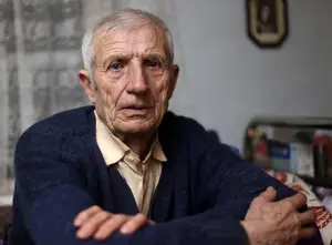 A photo of an elderly man sitting on a couch, looking to the side, with a serious expression and crossed arms, wearing a blue sweater and white shirt.