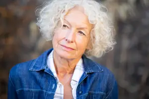 Portrait of an elderly woman with curly white hair wearing a denim jacket and a white shirt standing in a forest area