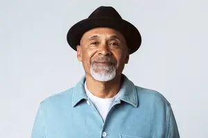 A smiling elderly man in a blue shirt and black hat posing for a photo against a white background.