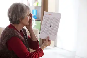 An elderly woman is sitting on a couch, holding a paper with a grid on it, and covering her left eye.