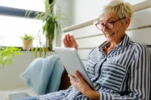 An elderly woman smiling while using a tablet in a room with plants and a window