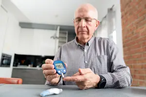 An elderly man is checking his blood sugar levels in the kitchen.