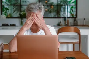 An elderly woman with gray hair is sitting in front of a table with a laptop, covering her eyes with her hands.
