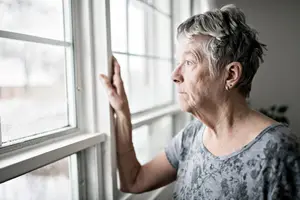 An elderly woman with short gray hair is looking outside the window, probably in a house.