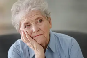 An elderly woman is sitting on a couch, looking at something with a sad expression on her face and her hand on her face.