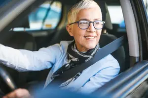 A smiling older woman wearing glasses and a scarf drives a car with her left hand on the steering wheel.