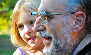 A young girl with blonde hair is standing next to an older man wearing glasses with a camera attached to them. The man is looking down at the girl and smiling, while the girl is looking up at him with her mouth open. They are standing in a grassy area with green trees in the background, and the man's glasses have a camera on the right side, attached to a metal frame.
