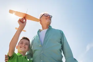 An older man wearing glasses is flying a wooden airplane with a boy on a sunny day