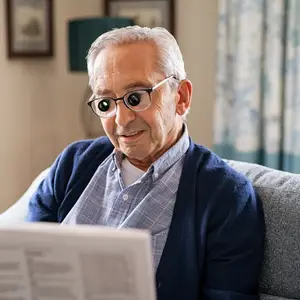 An elderly man with white hair and glasses is sitting on a couch and reading a newspaper.